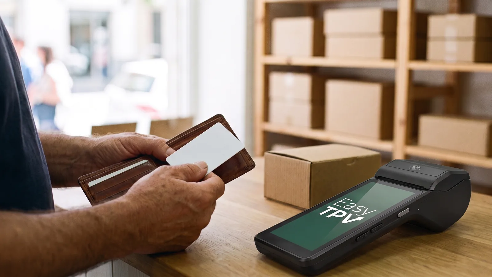 Mostrador de una tienda: las manos de un cliente sacan una tarjeta en blanco de la cartera, junto al datáfono y un platillo verde con monedas