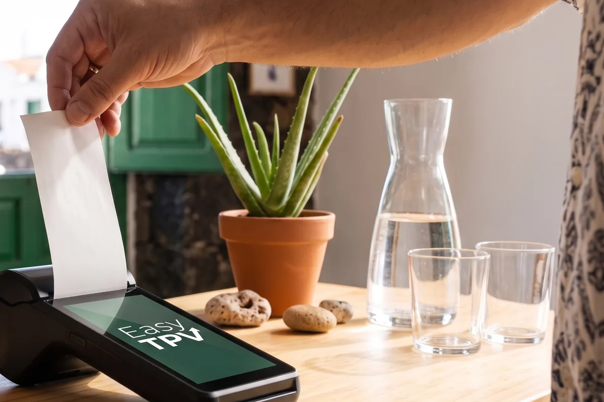 Mostrador de madera de un bar canario con una persiana verde y un aloe: la mano del dependiente corta el ticket que sale del datáfono